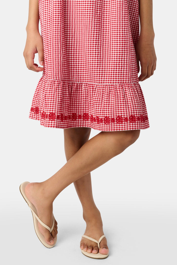 A young girl in the Roller Rabbit Kids Gingham Teagan Dress with an embroidered hem stands barefoot on a boat deck by the water, sunlight shimmering on the surface.