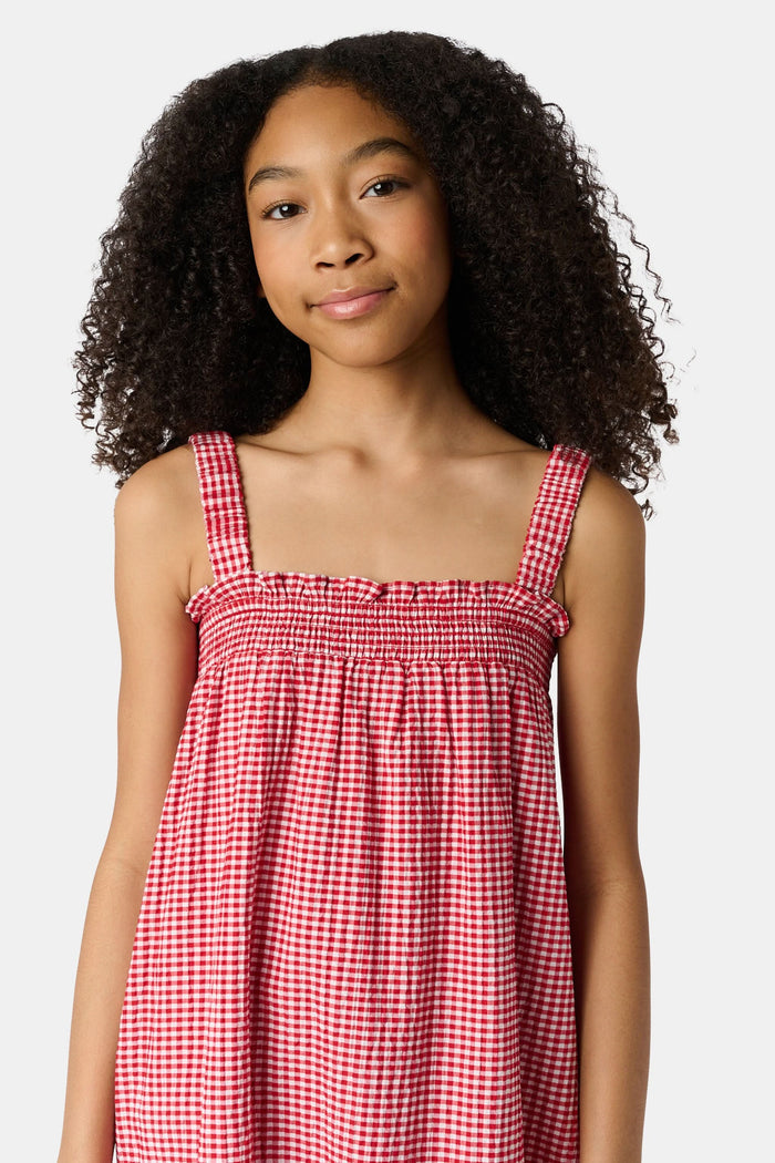 A young girl in the Roller Rabbit Kids Gingham Teagan Dress with an embroidered hem stands barefoot on a boat deck by the water, sunlight shimmering on the surface.