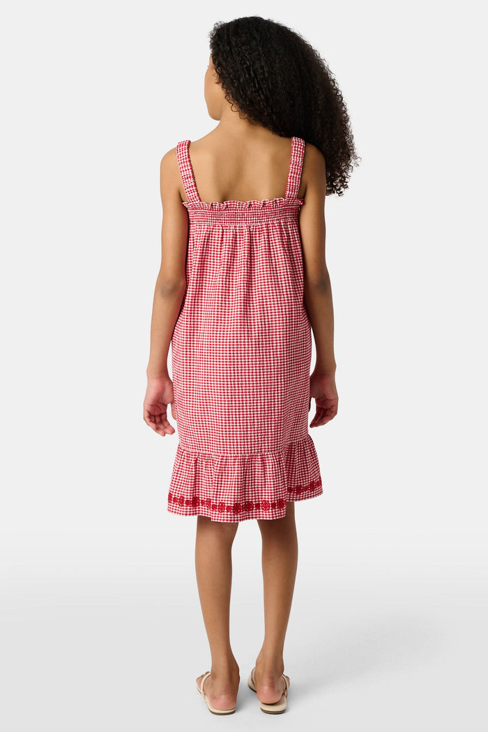 A young girl in the Roller Rabbit Kids Gingham Teagan Dress with an embroidered hem stands barefoot on a boat deck by the water, sunlight shimmering on the surface.