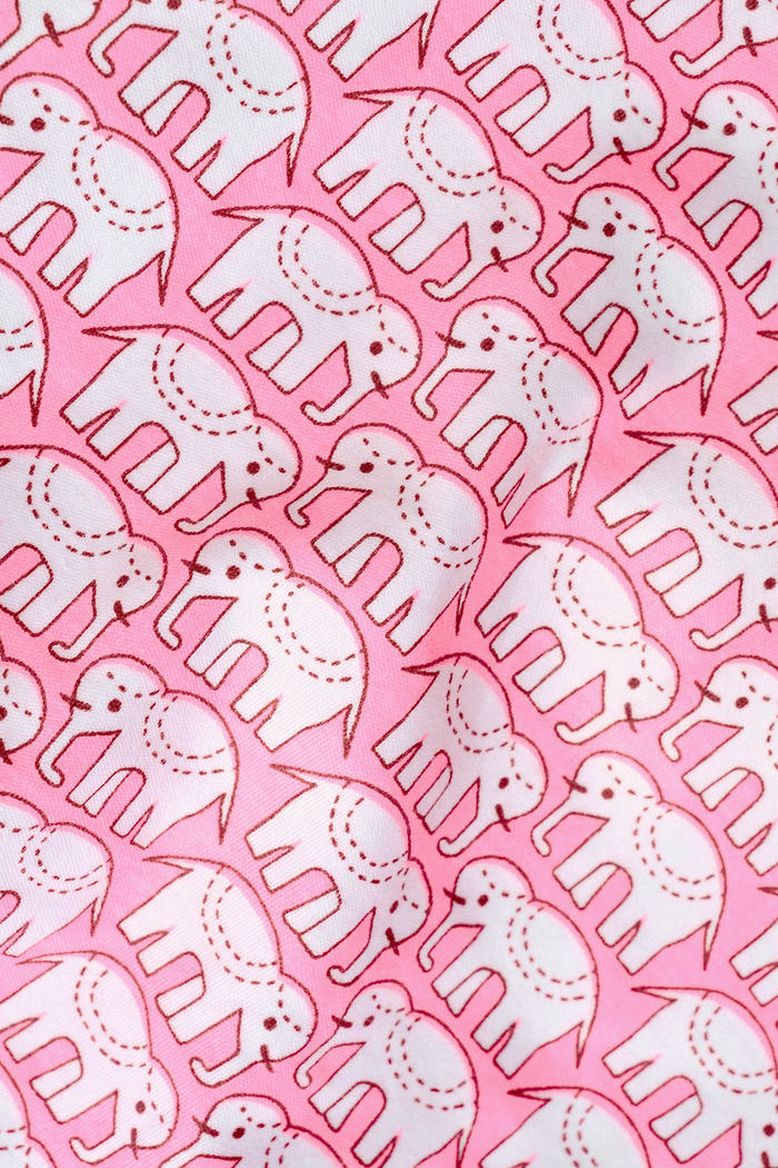 A woman stands barefoot, smiling, wearing Roller Rabbit's Hathi Long Sleeve Polo Pajamas—a pima cotton set in pink Hathi print with red piping and matching pants—one hand in her pocket against a plain light background.