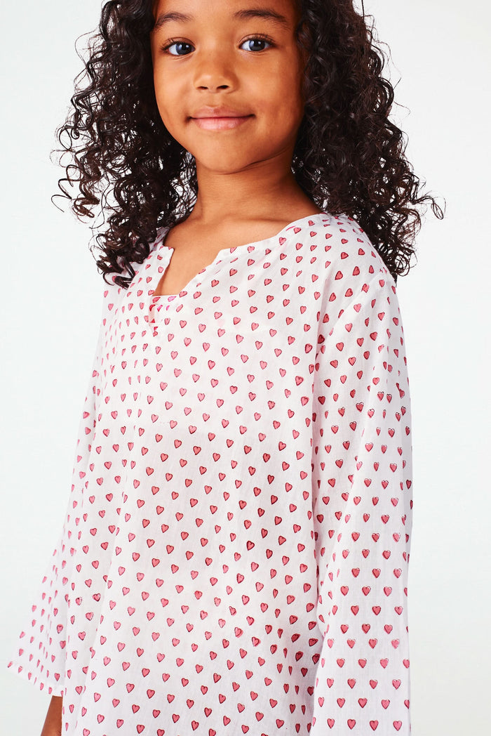 A young girl with curly hair smiles in a Roller Rabbit Kids Hearts Kurta, paired with light-colored shoes, standing against a plain white background.
