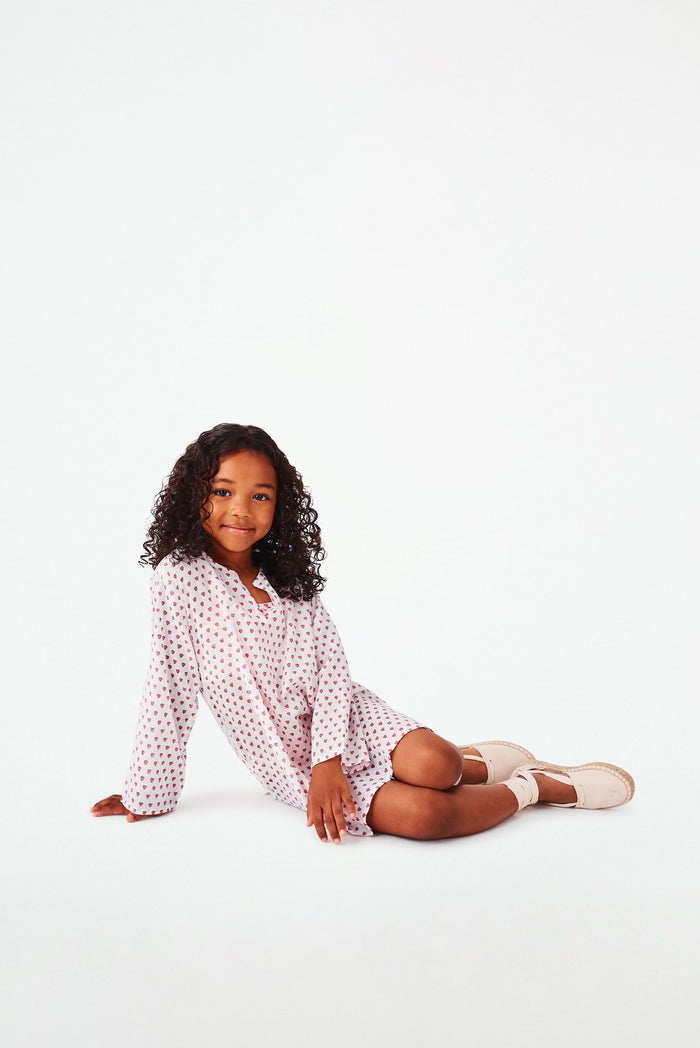 A young girl with curly hair smiles in a Roller Rabbit Kids Hearts Kurta, paired with light-colored shoes, standing against a plain white background.