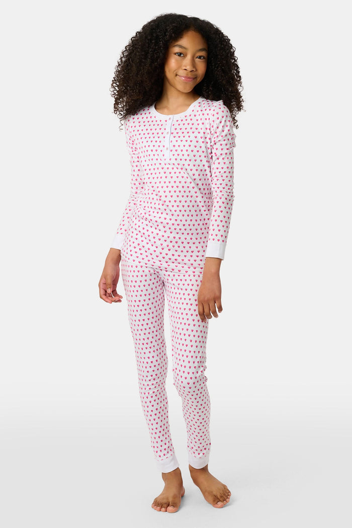 A young girl with curly hair stands barefoot, smiling slightly in Roller Rabbit’s Kids Hearts Pajamas—a premium cotton matching set—against a light gray background.