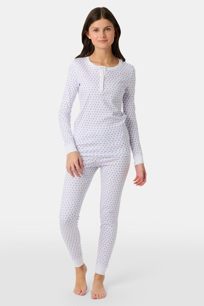 A girl stands barefoot in Roller Rabbit Kids Hearts Pajamas: a 100 cotton set with a fitted white long-sleeve top and purple heart-print pants with white cuffs, against a plain light background.