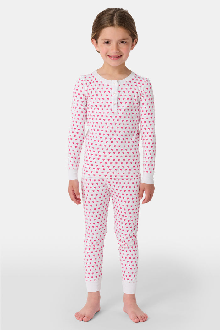 A young girl with curly hair stands barefoot, smiling slightly in Roller Rabbit’s Kids Hearts Pajamas—a premium cotton matching set—against a light gray background.