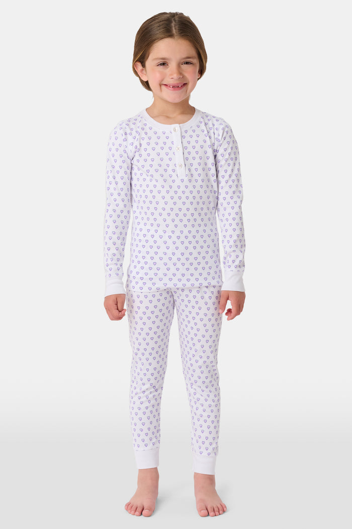 A girl stands barefoot in Roller Rabbit Kids Hearts Pajamas: a 100 cotton set with a fitted white long-sleeve top and purple heart-print pants with white cuffs, against a plain light background.