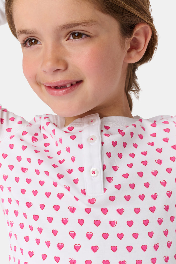 A young girl with curly hair stands barefoot, smiling slightly in Roller Rabbit’s Kids Hearts Pajamas—a premium cotton matching set—against a light gray background.