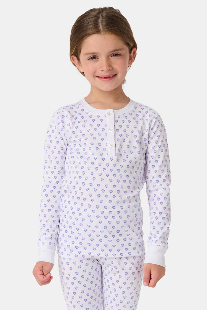 A girl stands barefoot in Roller Rabbit Kids Hearts Pajamas: a 100 cotton set with a fitted white long-sleeve top and purple heart-print pants with white cuffs, against a plain light background.
