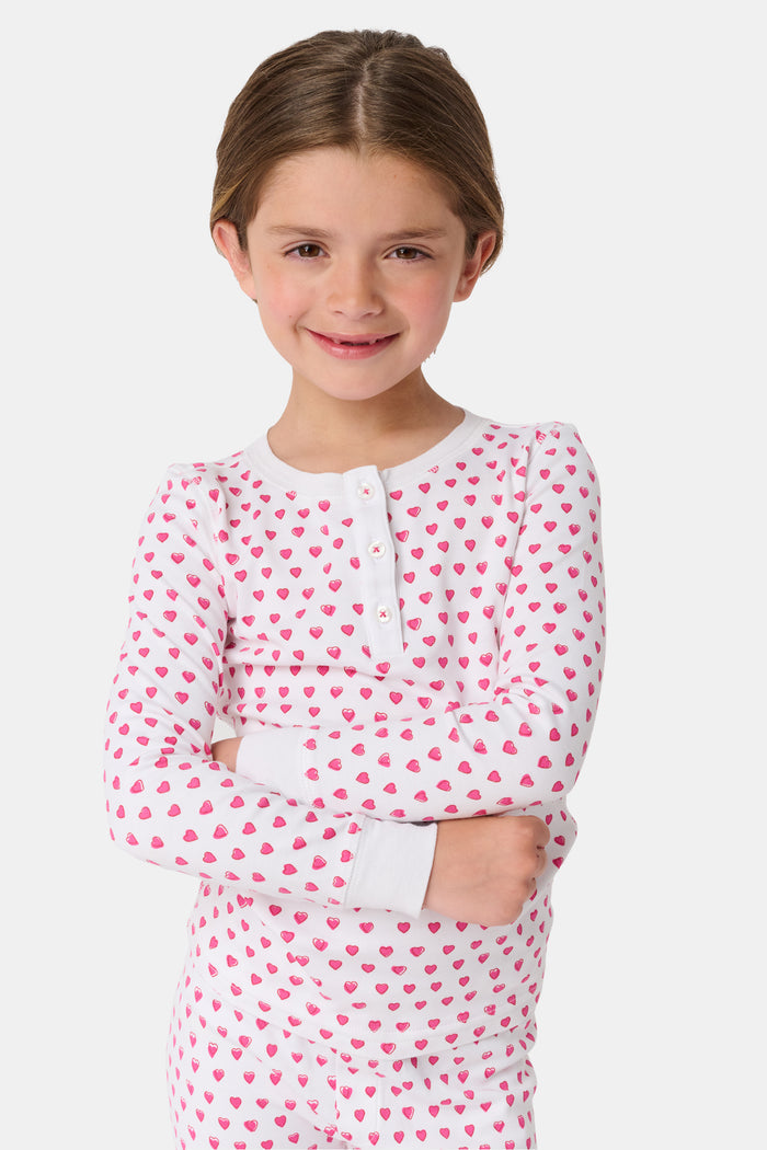 A young girl with curly hair stands barefoot, smiling slightly in Roller Rabbit’s Kids Hearts Pajamas—a premium cotton matching set—against a light gray background.