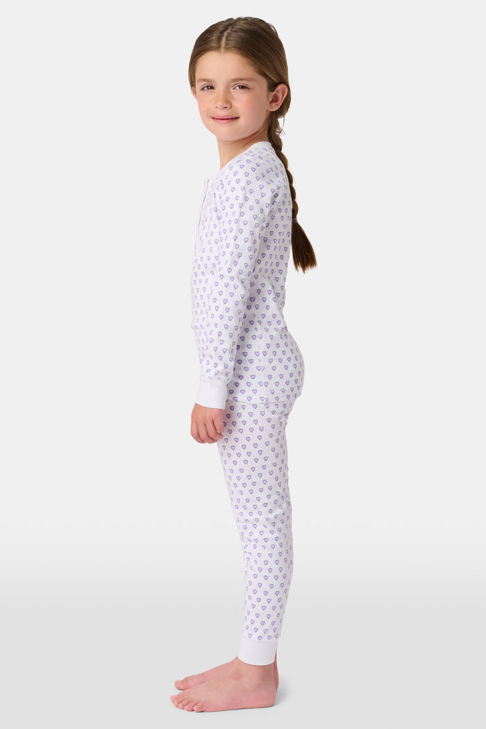 A girl stands barefoot in Roller Rabbit Kids Hearts Pajamas: a 100 cotton set with a fitted white long-sleeve top and purple heart-print pants with white cuffs, against a plain light background.