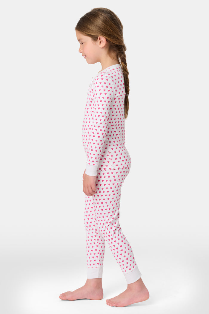 A young girl with curly hair stands barefoot, smiling slightly in Roller Rabbit’s Kids Hearts Pajamas—a premium cotton matching set—against a light gray background.