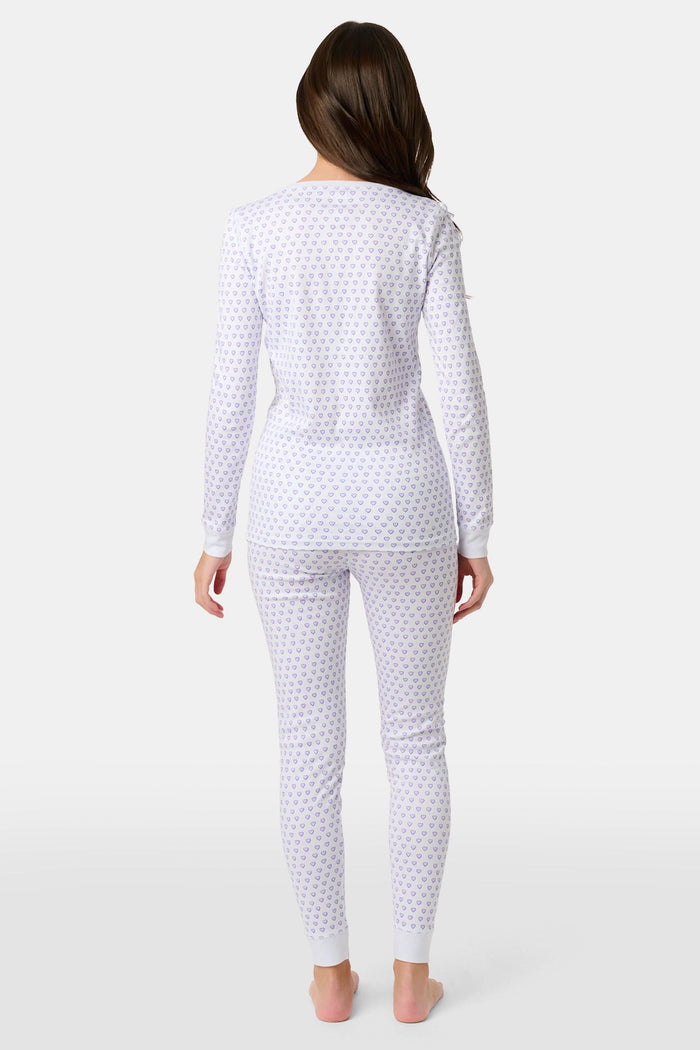 A girl stands barefoot in Roller Rabbit Kids Hearts Pajamas: a 100 cotton set with a fitted white long-sleeve top and purple heart-print pants with white cuffs, against a plain light background.