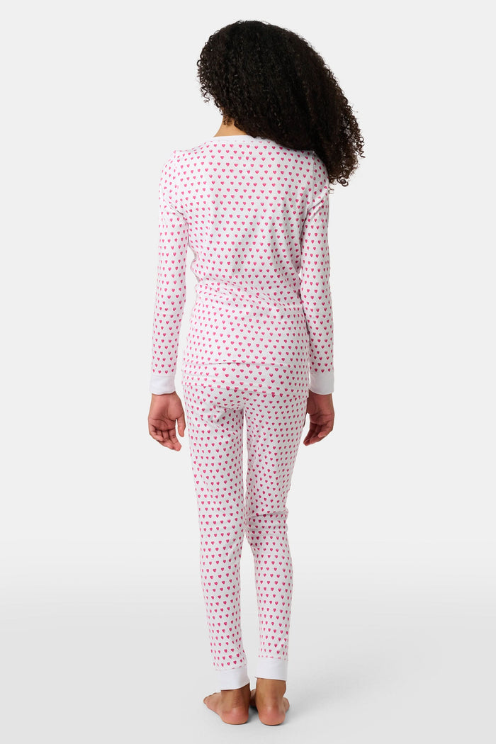 A young girl with curly hair stands barefoot, smiling slightly in Roller Rabbit’s Kids Hearts Pajamas—a premium cotton matching set—against a light gray background.