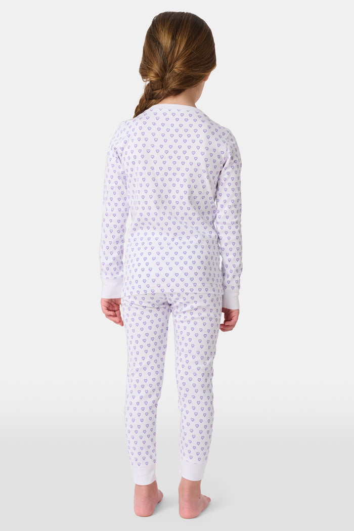 A girl stands barefoot in Roller Rabbit Kids Hearts Pajamas: a 100 cotton set with a fitted white long-sleeve top and purple heart-print pants with white cuffs, against a plain light background.
