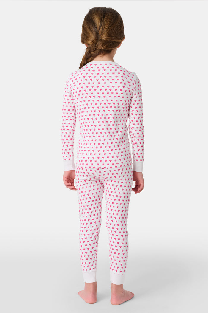 A young girl with curly hair stands barefoot, smiling slightly in Roller Rabbit’s Kids Hearts Pajamas—a premium cotton matching set—against a light gray background.