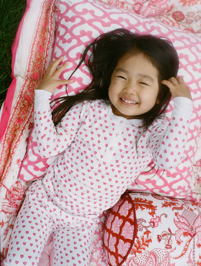 A young girl with curly hair stands barefoot, smiling slightly in Roller Rabbit’s Kids Hearts Pajamas—a premium cotton matching set—against a light gray background.