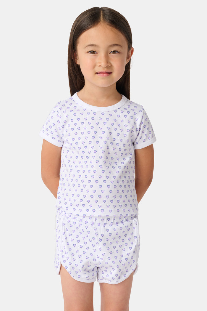 A young child with long brown hair stands facing forward, smiling with hands behind their back. They wear the Roller Rabbit Kids Hearts Playa Play Set. The background is plain white.