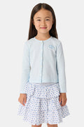 A young girl with long brown hair wears a light blue Roller Rabbit Kids Hearts Pointelle Heidi Cardigan over a white tiered skirt with blue heart patterns, standing and smiling against a plain white background.