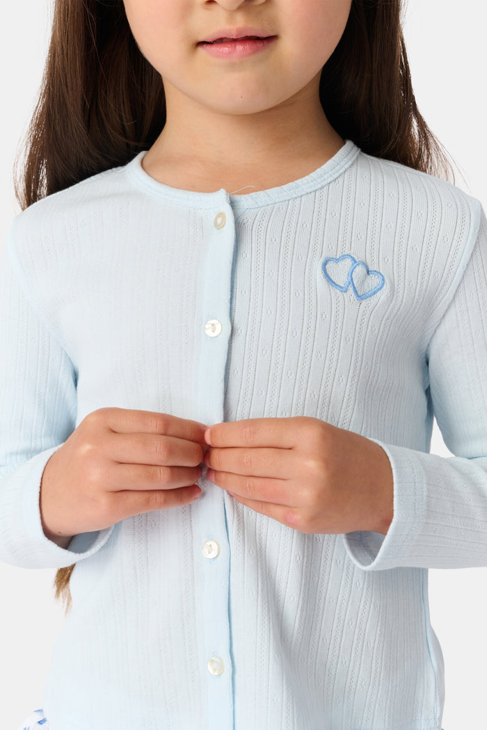 A young girl with long brown hair wears a light blue Roller Rabbit Kids Hearts Pointelle Heidi Cardigan over a white tiered skirt with blue heart patterns, standing and smiling against a plain white background.