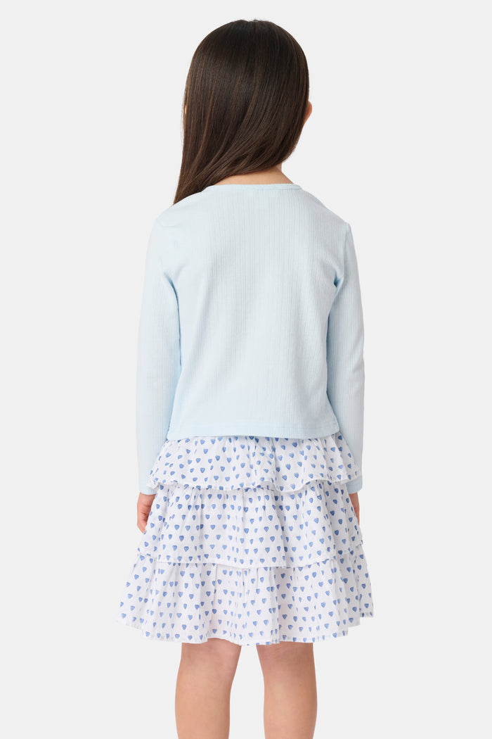 A young girl with long brown hair wears a light blue Roller Rabbit Kids Hearts Pointelle Heidi Cardigan over a white tiered skirt with blue heart patterns, standing and smiling against a plain white background.