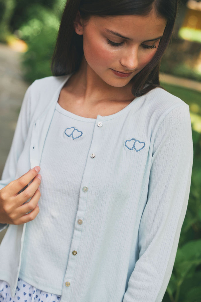 A young girl with long brown hair wears a light blue Roller Rabbit Kids Hearts Pointelle Heidi Cardigan over a white tiered skirt with blue heart patterns, standing and smiling against a plain white background.