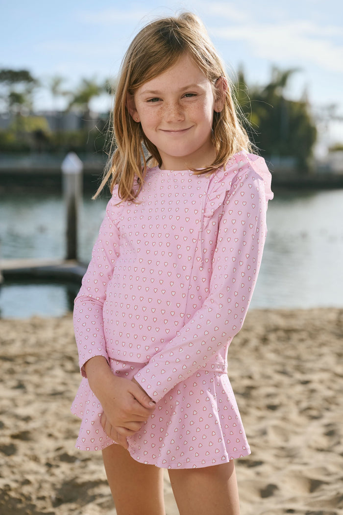 A young girl smiles on a sandy beach, wearing the Roller Rabbit Kids Hearts Ruffle Rashguard with long sleeves in light pink. Water and trees blur in the background, showcasing stylish kids swimwear for sunny days.