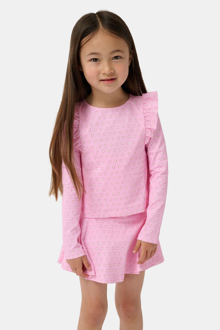 A young girl smiles on a sandy beach, wearing the Roller Rabbit Kids Hearts Ruffle Rashguard with long sleeves in light pink. Water and trees blur in the background, showcasing stylish kids swimwear for sunny days.