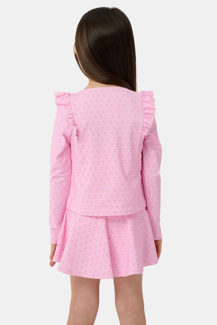 A young girl smiles on a sandy beach, wearing the Roller Rabbit Kids Hearts Ruffle Rashguard with long sleeves in light pink. Water and trees blur in the background, showcasing stylish kids swimwear for sunny days.