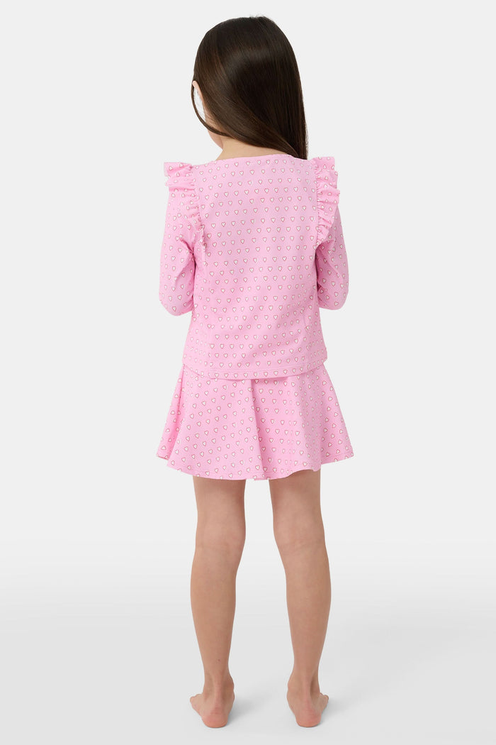 A young girl smiles on a sandy beach, wearing the Roller Rabbit Kids Hearts Ruffle Rashguard with long sleeves in light pink. Water and trees blur in the background, showcasing stylish kids swimwear for sunny days.