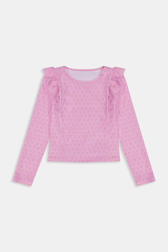 A young girl smiles on a sandy beach, wearing the Roller Rabbit Kids Hearts Ruffle Rashguard with long sleeves in light pink. Water and trees blur in the background, showcasing stylish kids swimwear for sunny days.