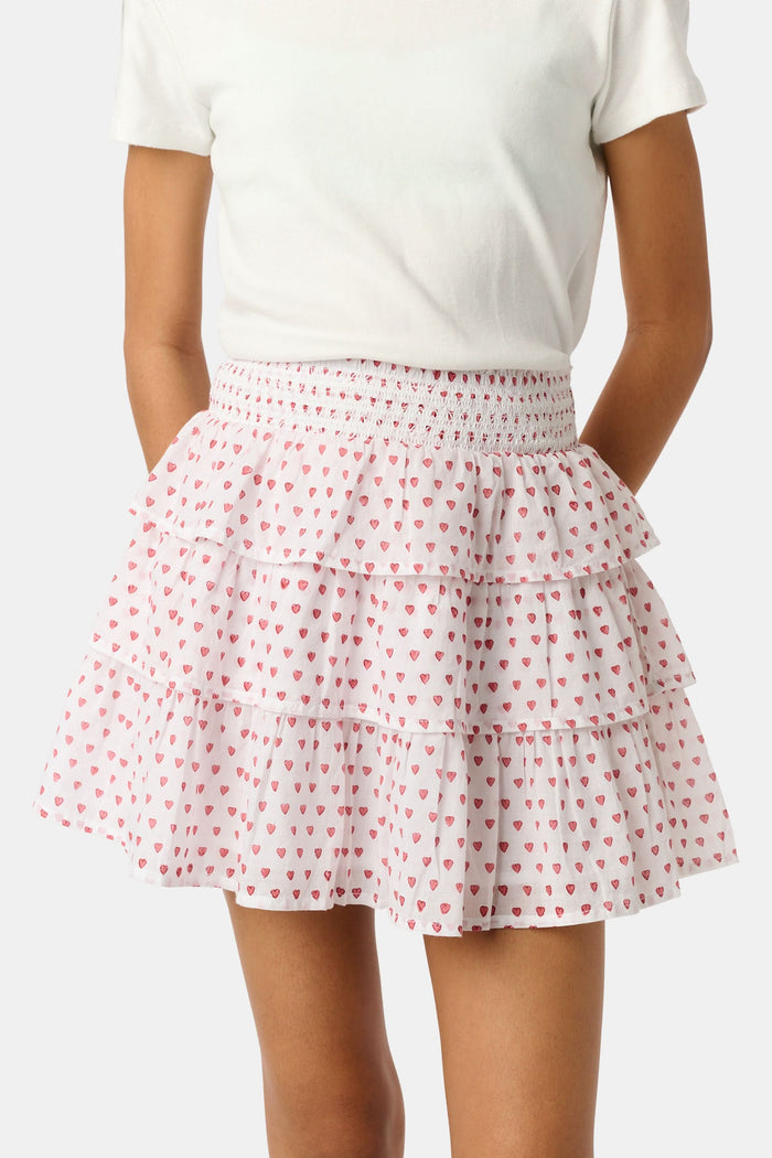 A person is wearing a white short-sleeve top and the Roller Rabbit Kids Hearts Sami Skirt, which features pink hearts, standing with hands behind their back against a plain light background.