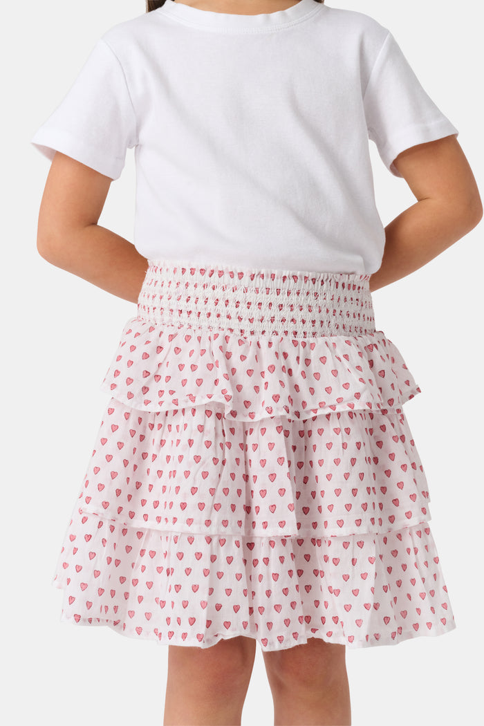 A person is wearing a white short-sleeve top and the Roller Rabbit Kids Hearts Sami Skirt, which features pink hearts, standing with hands behind their back against a plain light background.