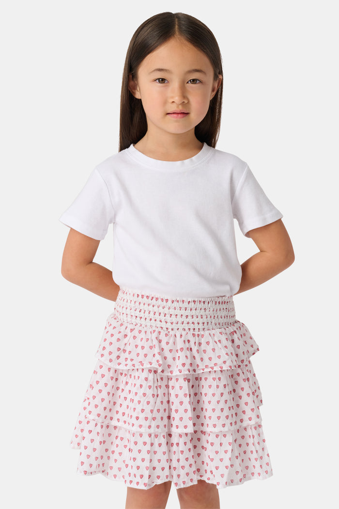 A person is wearing a white short-sleeve top and the Roller Rabbit Kids Hearts Sami Skirt, which features pink hearts, standing with hands behind their back against a plain light background.