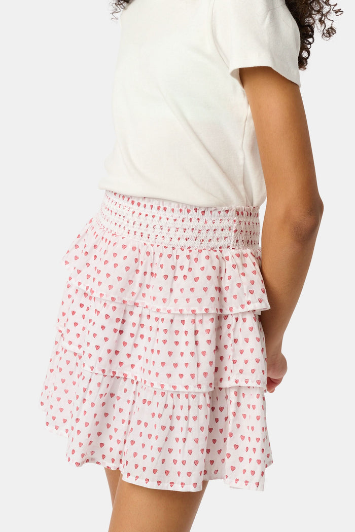 A person is wearing a white short-sleeve top and the Roller Rabbit Kids Hearts Sami Skirt, which features pink hearts, standing with hands behind their back against a plain light background.