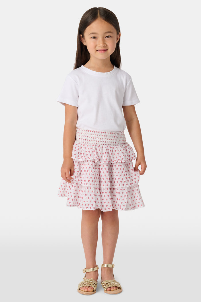 A person is wearing a white short-sleeve top and the Roller Rabbit Kids Hearts Sami Skirt, which features pink hearts, standing with hands behind their back against a plain light background.