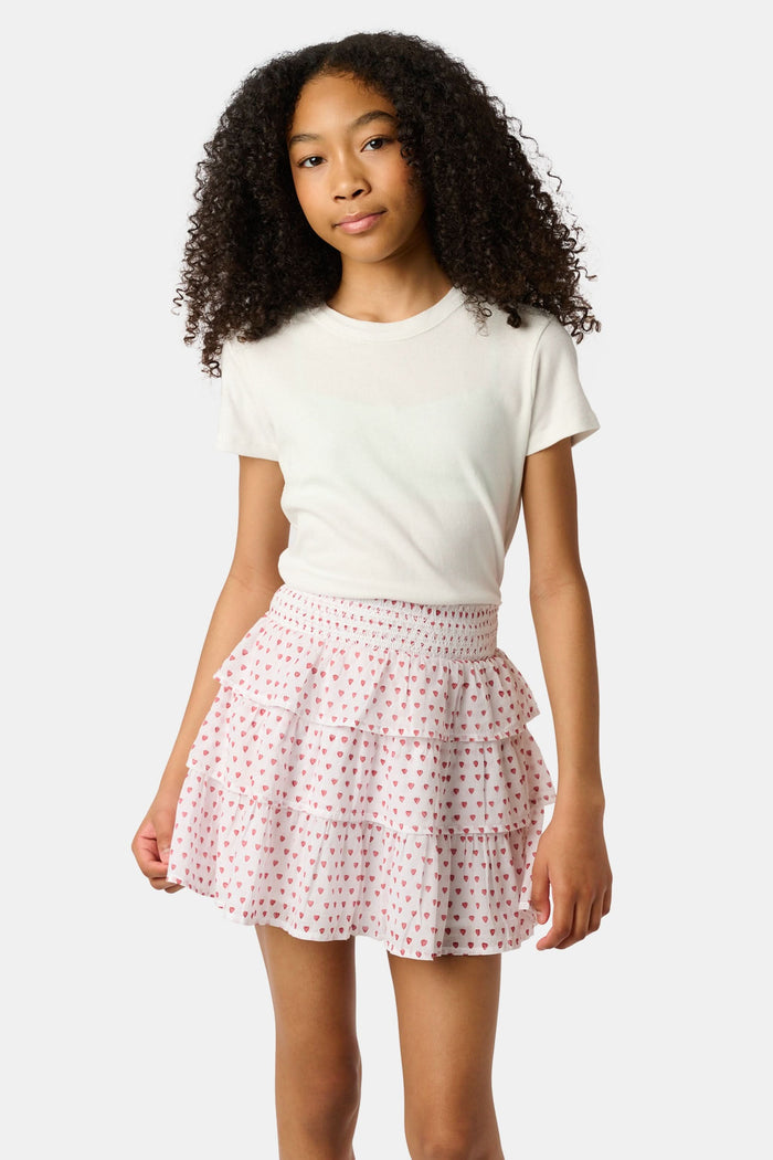 A person is wearing a white short-sleeve top and the Roller Rabbit Kids Hearts Sami Skirt, which features pink hearts, standing with hands behind their back against a plain light background.