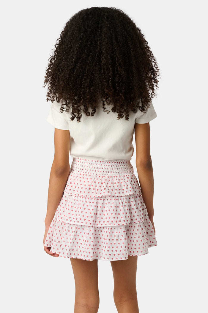 A person is wearing a white short-sleeve top and the Roller Rabbit Kids Hearts Sami Skirt, which features pink hearts, standing with hands behind their back against a plain light background.