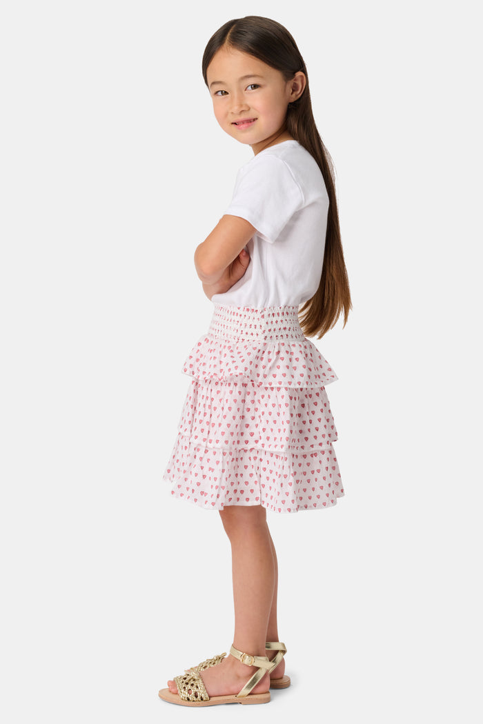 A person is wearing a white short-sleeve top and the Roller Rabbit Kids Hearts Sami Skirt, which features pink hearts, standing with hands behind their back against a plain light background.