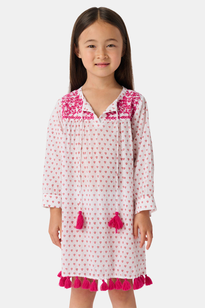 A young girl with long brown hair smiles in the Roller Rabbit Kids Hearts Serafina Tunic, a cotton dress with pink heart patterns, chest embroidery, and pink tassels at the neckline and hem, standing against a plain light background.