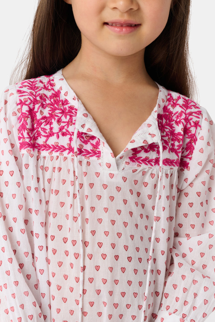 A young girl with long brown hair smiles in the Roller Rabbit Kids Hearts Serafina Tunic, a cotton dress with pink heart patterns, chest embroidery, and pink tassels at the neckline and hem, standing against a plain light background.