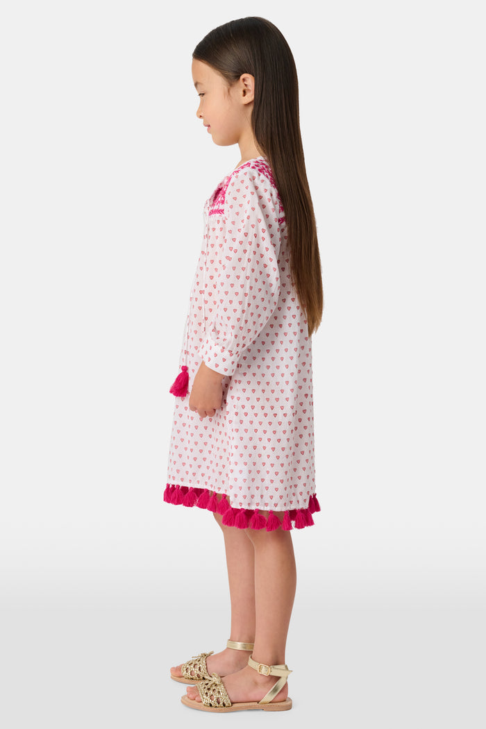 A young girl with long brown hair smiles in the Roller Rabbit Kids Hearts Serafina Tunic, a cotton dress with pink heart patterns, chest embroidery, and pink tassels at the neckline and hem, standing against a plain light background.