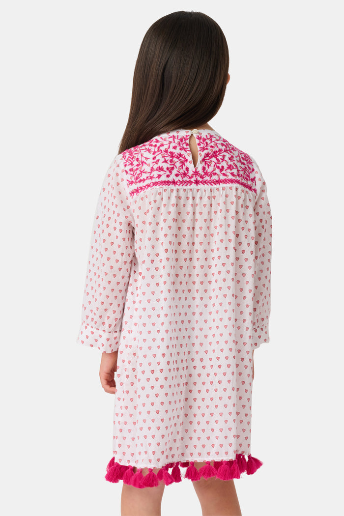 A young girl with long brown hair smiles in the Roller Rabbit Kids Hearts Serafina Tunic, a cotton dress with pink heart patterns, chest embroidery, and pink tassels at the neckline and hem, standing against a plain light background.