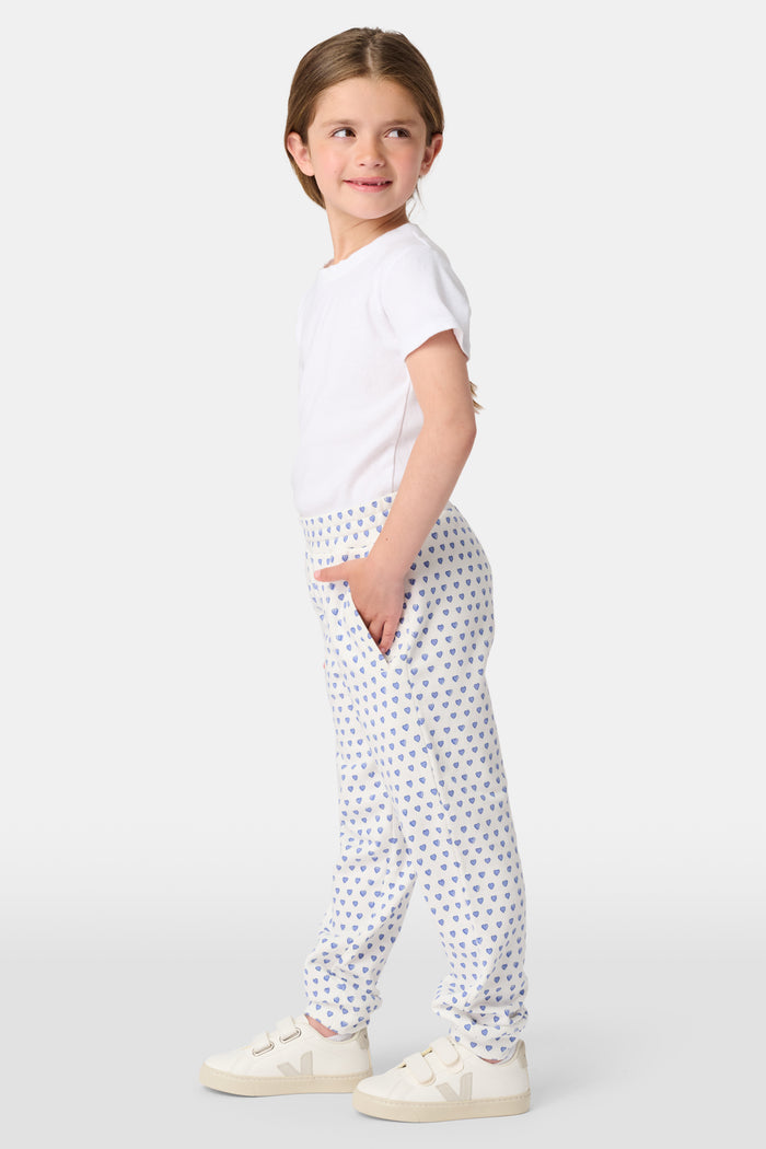 A young child smiles while wearing Roller Rabbit Kids Hearts Sweatpants and a plain white t-shirt, with one hand in the pocket and looking slightly to the side.