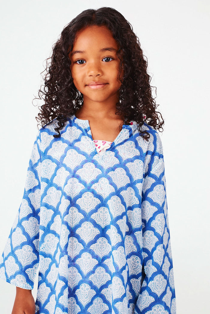 A young girl with curly hair smiles against a white background, wearing the Roller Rabbit Kids Jemina Kurta in blue and white cotton, paired with white espadrille sandals.