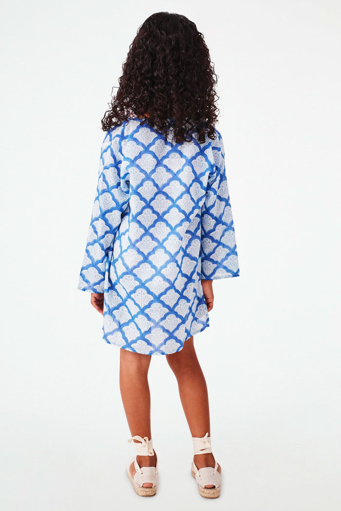 A young girl with curly hair smiles against a white background, wearing the Roller Rabbit Kids Jemina Kurta in blue and white cotton, paired with white espadrille sandals.