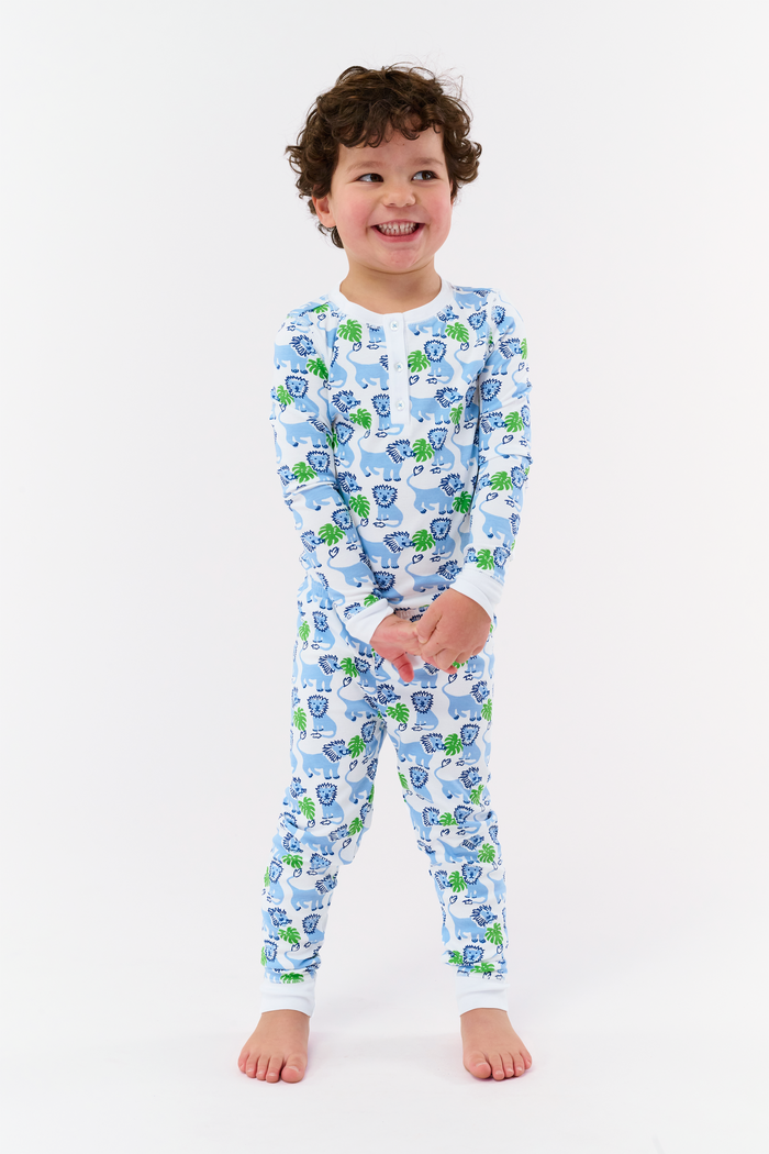 A young child with curly hair stands barefoot, smiling in Roller Rabbit Kids Leo The Lion Pajamas made from soft Pima cotton, against a plain white background.