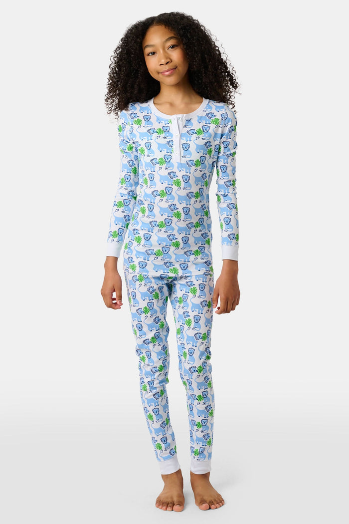 A young child with curly hair stands barefoot, smiling in Roller Rabbit Kids Leo The Lion Pajamas made from soft Pima cotton, against a plain white background.