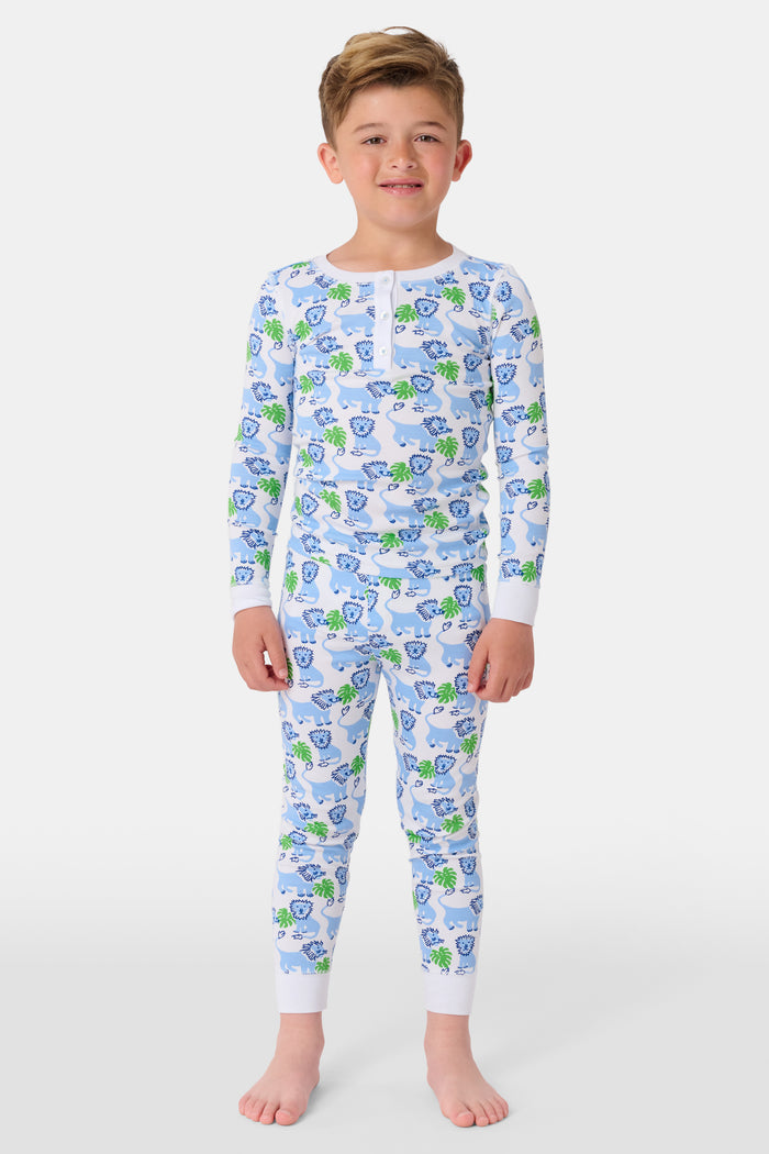 A young child with curly hair stands barefoot, smiling in Roller Rabbit Kids Leo The Lion Pajamas made from soft Pima cotton, against a plain white background.