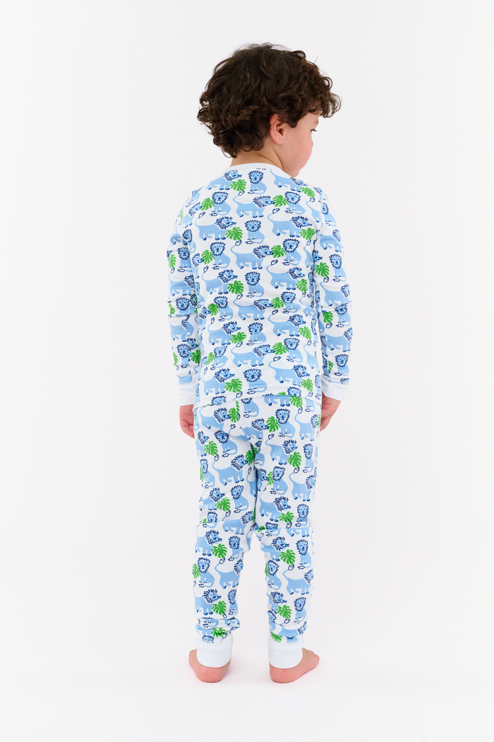 A young child with curly hair stands barefoot, smiling in Roller Rabbit Kids Leo The Lion Pajamas made from soft Pima cotton, against a plain white background.