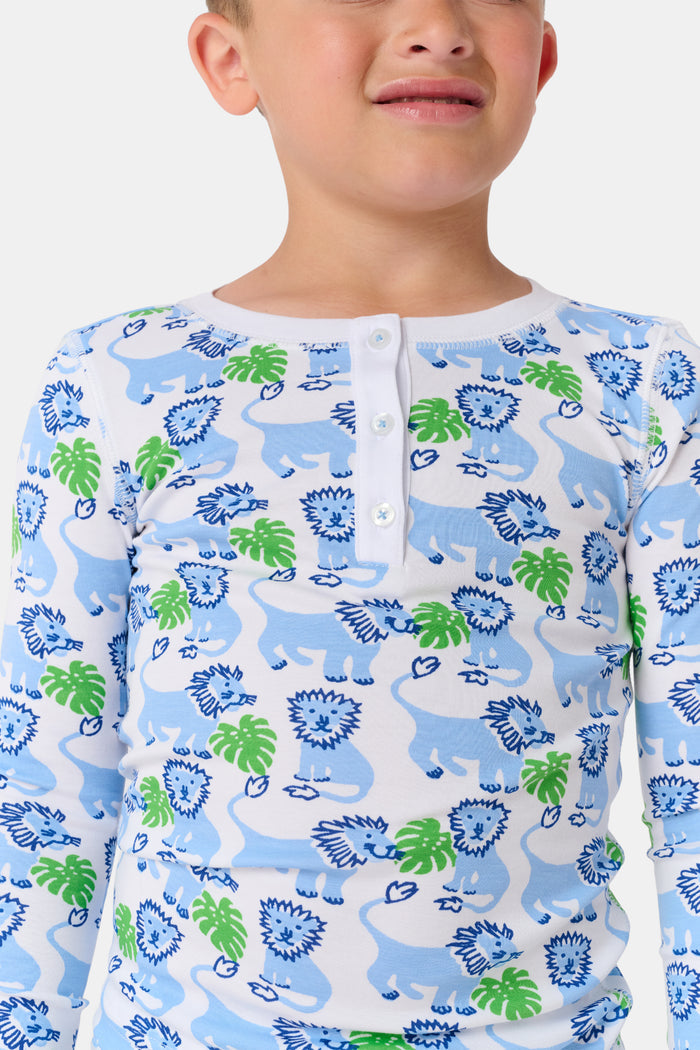 A young child with curly hair stands barefoot, smiling in Roller Rabbit Kids Leo The Lion Pajamas made from soft Pima cotton, against a plain white background.
